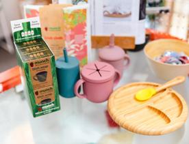 A range of Munch Cupboard products sit on a table, including a bamboo plate and spoon, and silicon drinking cups.