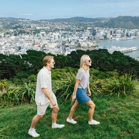 Two people walking on lush green grass on top of Mount Victoria. Wellington city and harbour are in the background, surrounded by green hills in the distance.