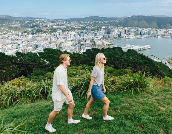 Two people walking on lush green grass on top of Mount Victoria. Wellington city and harbour are in the background, surrounded by green hills in the distance.