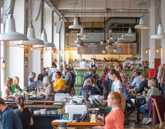 The busy and vibrant interior of Charley Noble, with customers sitting at tables and servers waiting tables, and kitchen staff in the background preparing food.