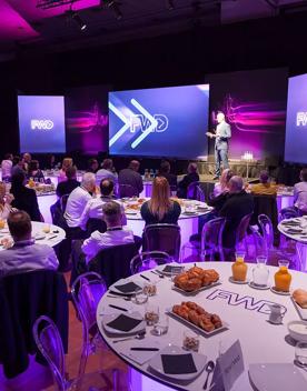 Inside Shed 6 at a private function, with a stage set up, tables and chairs and pink lights shining down.