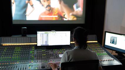 A person sits at a sound desk with one small screen and one large screen in front of them.