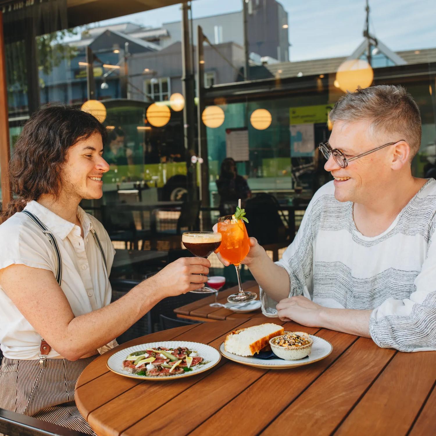 Two people cheers their cocktails and enjoy a couple sharing plates on an outdoor patio.