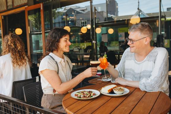 Two people cheers their cocktails and enjoy a couple sharing plates on an outdoor patio.