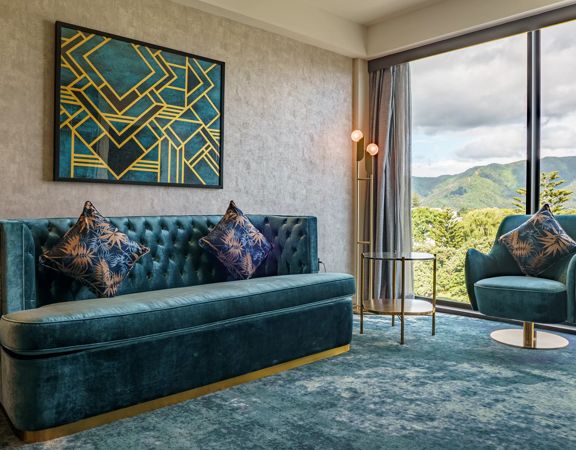 A room inside The Sebel in Hutt Valley, theresd a alrge couch and small chair both a velvet teal colour with gold accents.