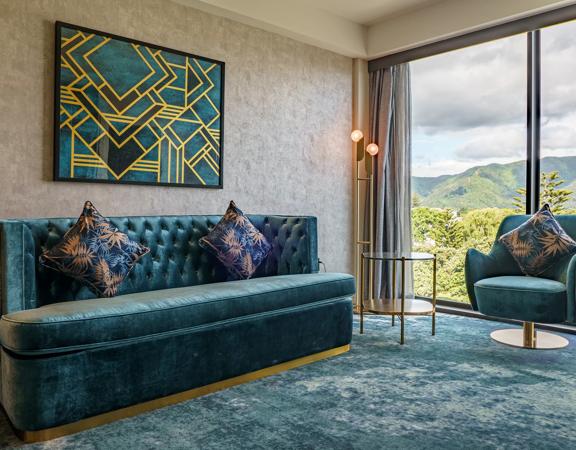 A room inside The Sebel in Hutt Valley, theresd a alrge couch and small chair both a velvet teal colour with gold accents.