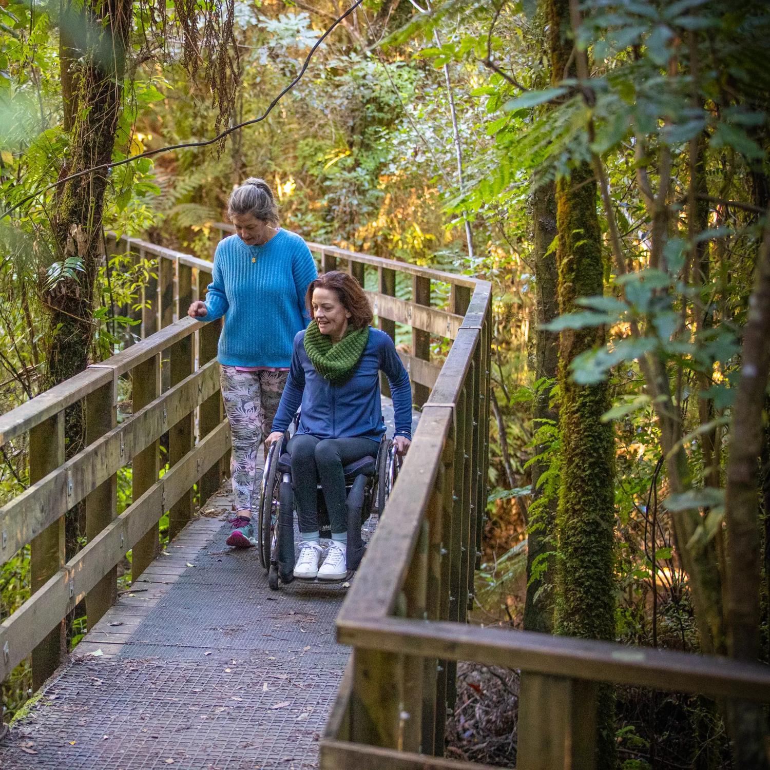 Two people are on a nature trail going across a bridge/boardwalk. One is a wheelchair user.