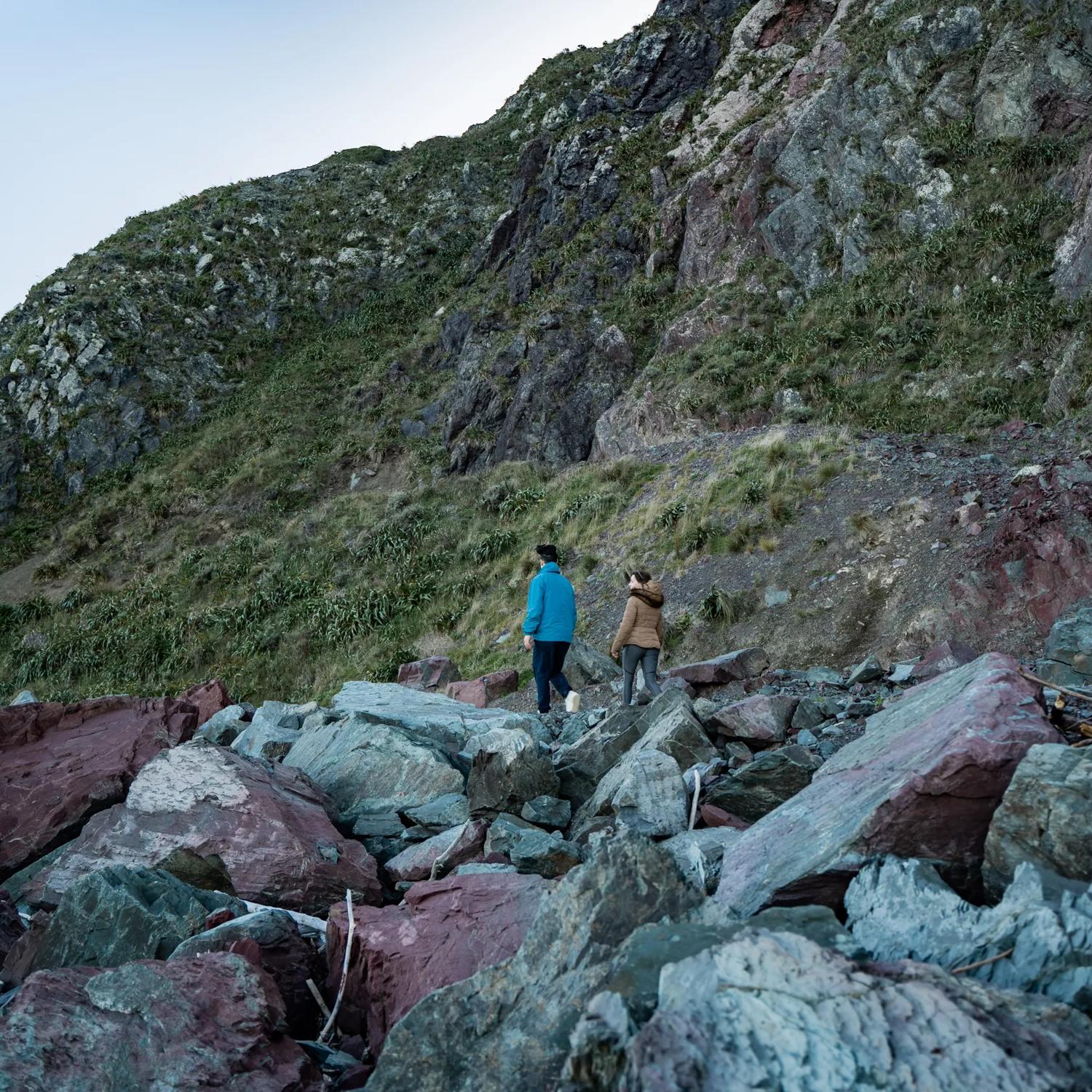 2 people walk along large rocks, on the Red Rocks Coastal Walkway.