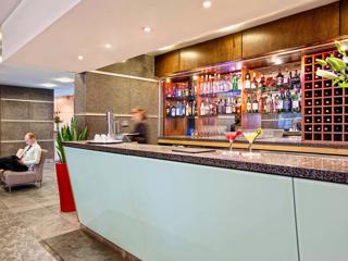 A hotel bar counter with two cocktails in martini glasses, a worker walking, and shelves with liquor bottles are on display.