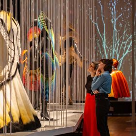 Museum visitors view a display of flowing garments and costume pieces behind hanging vertical strings in a gallery exhibition.