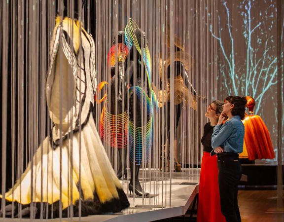 Museum visitors view a display of flowing garments and costume pieces behind hanging vertical strings in a gallery exhibition.