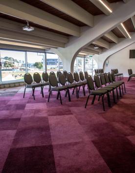 The Longroom in the grandstand at the Basin Reserve. Three rows of chairs sit in front of a projector screen with the back of the room looking out on the cricket pitch.