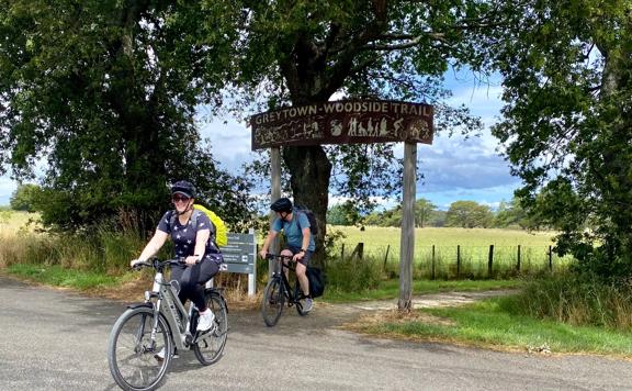 Two cyclists ride past the Greytown to Woodside Trail entrance sign.