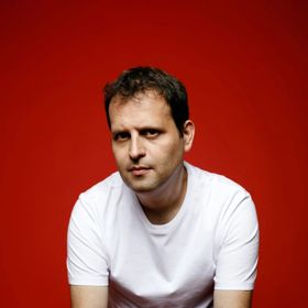 Adam Kay seated on a stool against a red background, wearing a white T-shirt and dark jeans, with hands resting in front.