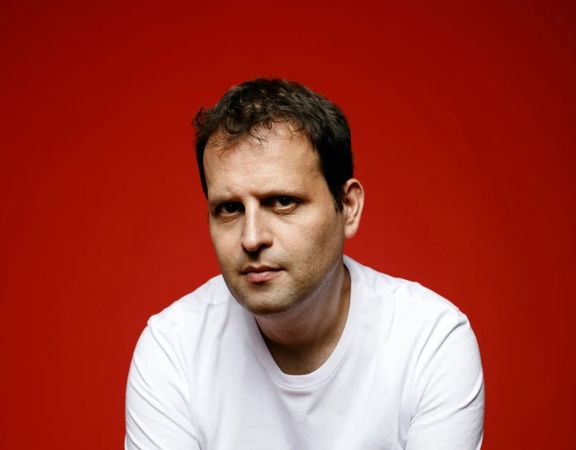 Adam Kay seated on a stool against a red background, wearing a white T-shirt and dark jeans, with hands resting in front.