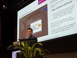 A presenter stands at a podium speaking to an audience, with a large projected slide showing code and documentation behind them.