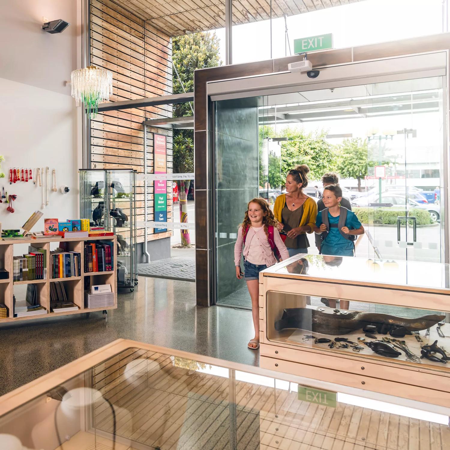 A family of three walking in to the foyer at Aratoi. The gift shop can be seen to the left of the image. The foyer has large modern glass windows and wooden slats near the entrance doors.