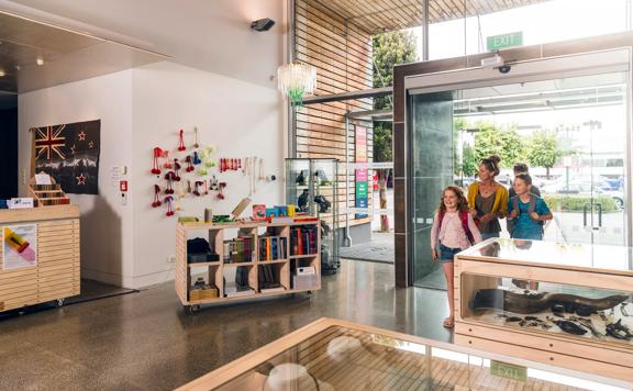 A family of three walking in to the foyer at Aratoi. The gift shop can be seen to the left of the image. The foyer has large modern glass windows and wooden slats near the entrance doors.