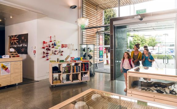 A family of three walking in to the foyer at Aratoi. The gift shop can be seen to the left of the image. The foyer has large modern glass windows and wooden slats near the entrance doors.