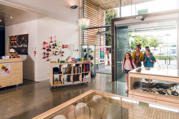 A family of three walking in to the foyer at Aratoi. The gift shop can be seen to the left of the image. The foyer has large modern glass windows and wooden slats near the entrance doors.