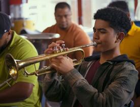 A still from the film Red White and Brass with a person wearing a grey and brown plaid shirt playing the trumpet and three other musicians siting behind him.