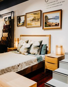 Bedroom showroom display at YOYO Design by Kiwi in Wellington, featuring a wooden bed with patterned cushions, bedside tables, framed landscape artwork, and homewares arranged against a white wall.