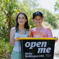 Two smiling people hold a black plastic box with a yellow lid while standing on a nature trail.