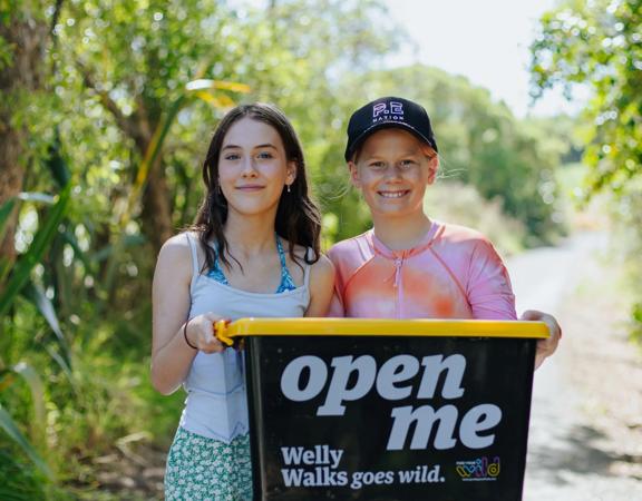 Two smiling people hold a black plastic box with a yellow lid while standing on a nature trail.