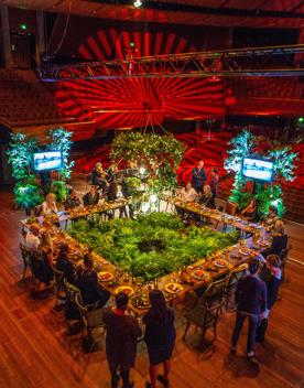 A stage set up at the Michael Fowler Centre in Wellington. People surround a tables that have been arranged into a square with space in the centre for green foliage.