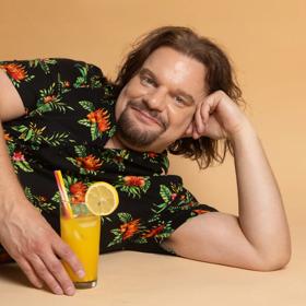 Finnish comedian, Ismo Leikola, wearing a black floral Hawaiian shirt, lying on his left side with an orange drink.