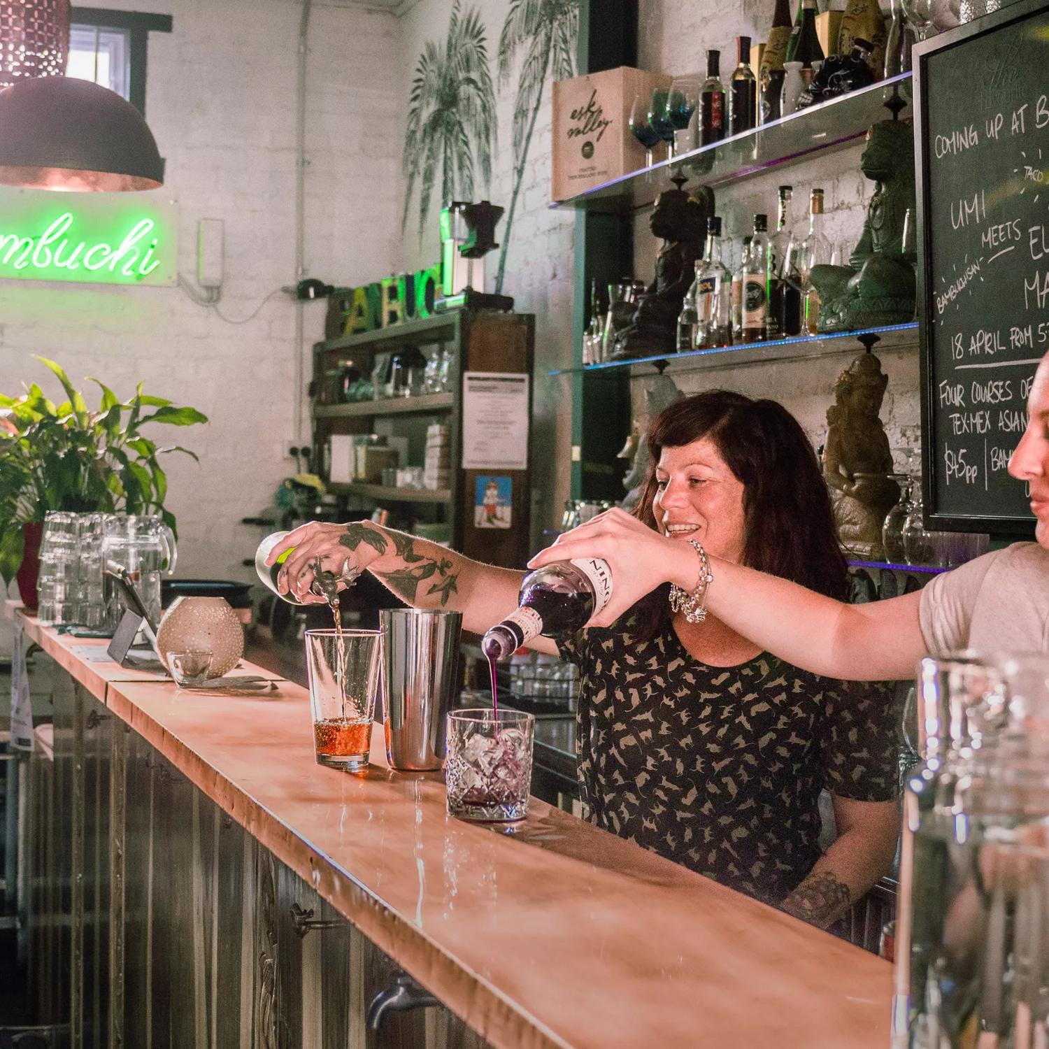 Two bartenders pouring drinks at the BambuchiSan bar, Green indoor plants occupy the background.