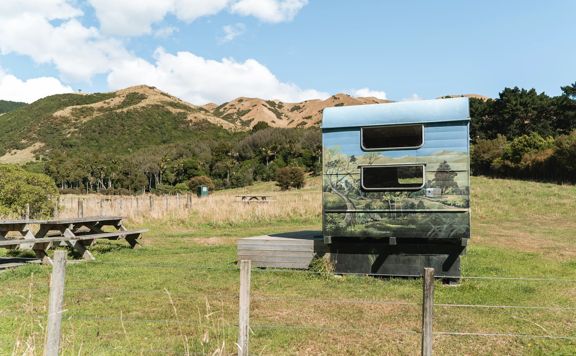 A caravan with a beach scene painted on it sits in the middle of a farm paddock on Whareroa Farm.