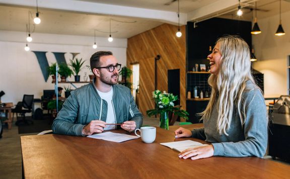 Two people sit at a wooden table in an industrial-style office. They're talking and have paper in front of them.