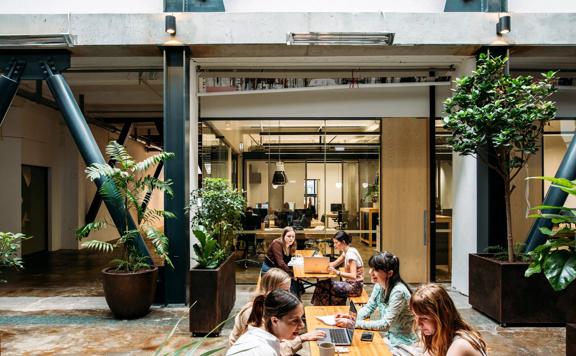 People sit at the wooden tables inside The Atrium at The Exchange. There are large planters surrounding them.