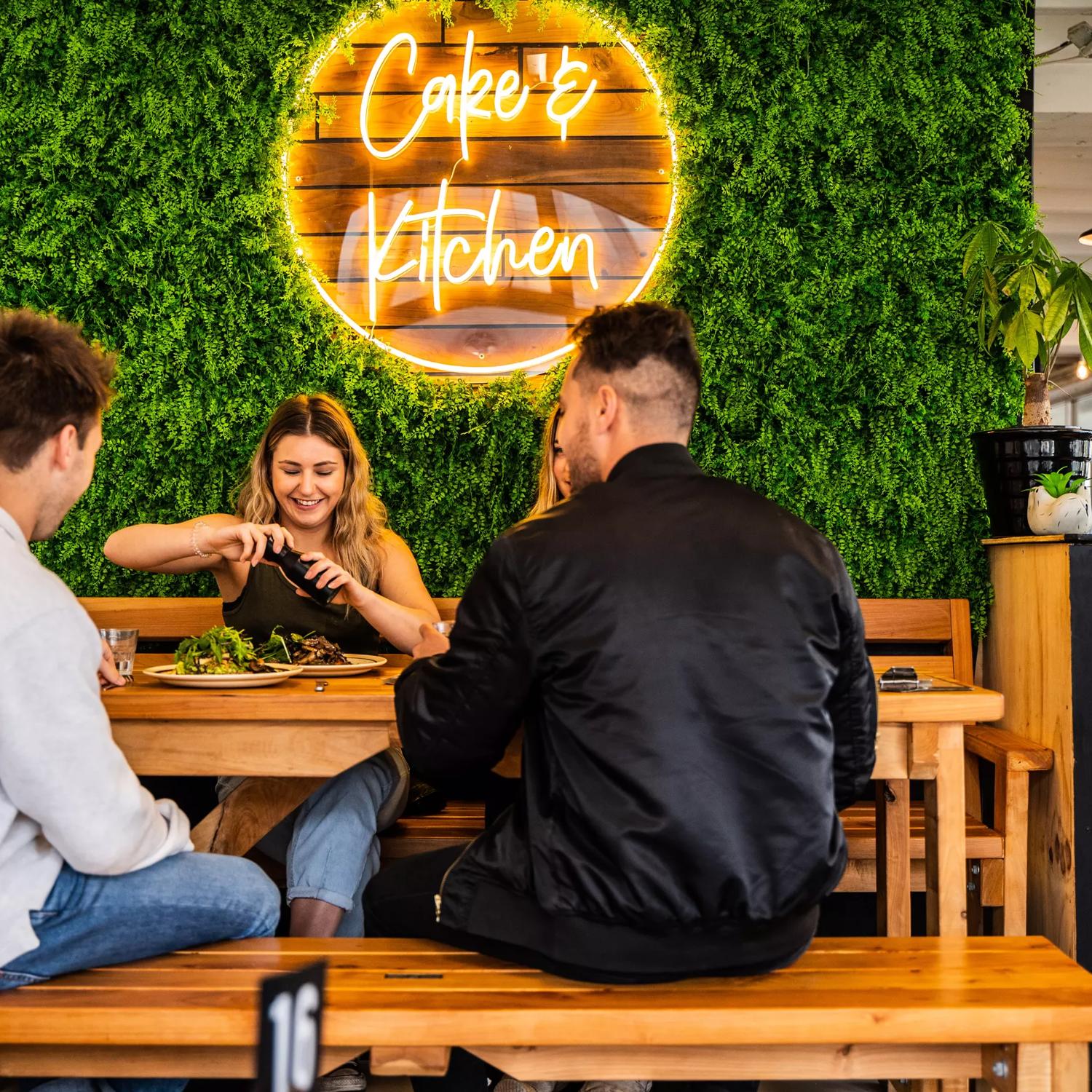 A group of people having food in front of a plant wall with a luminescent sign saying Cake & Kitchen.