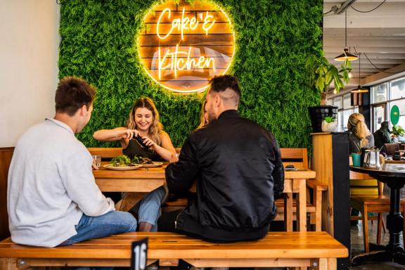 A group of people having food in front of a plant wall with a luminescent sign saying Cake & Kitchen.