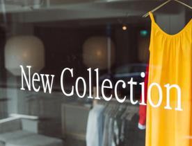 The front window of a boutique clothing store that reads "New Collection" with a bright yellow dress on display.