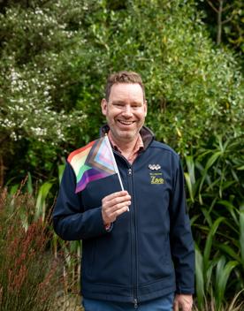 A Wellington Zoo employee stands, smiles and waves a LGBTQIA+ flag in his right hand in front of green bushes and trees.