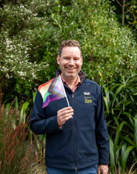 A Wellington Zoo employee stands, smiles and waves a LGBTQIA+ flag in his right hand in front of green bushes and trees. 