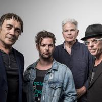 The members of Dragon, a New Zealand rock band, are standing together in front of a light grey background. 
