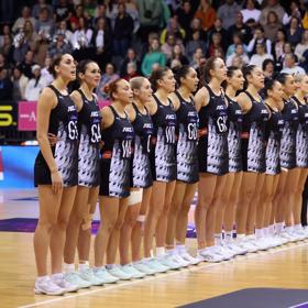The Silver Ferns, New Zealand national netball team, stand in a row on the court with fans in the stands.