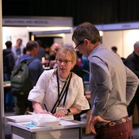 2 people talking over a table while the busy background of a convention goes on behind them.