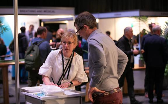 2 people talking over a table while the busy background of a convention goes on behind them.