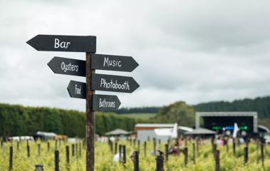 A sign post pointing to the bar, oysters, food, music, photobooth and bathrooms at an outdoor festival.