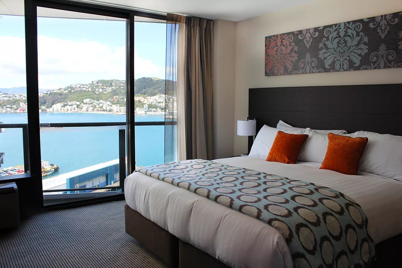 A room at the Rydges Hotel located at 75 Featherston Street, Pipitea, Wellington, with a king-sized bed, grey carpet, beige walls and a large window with a view of the harbour.