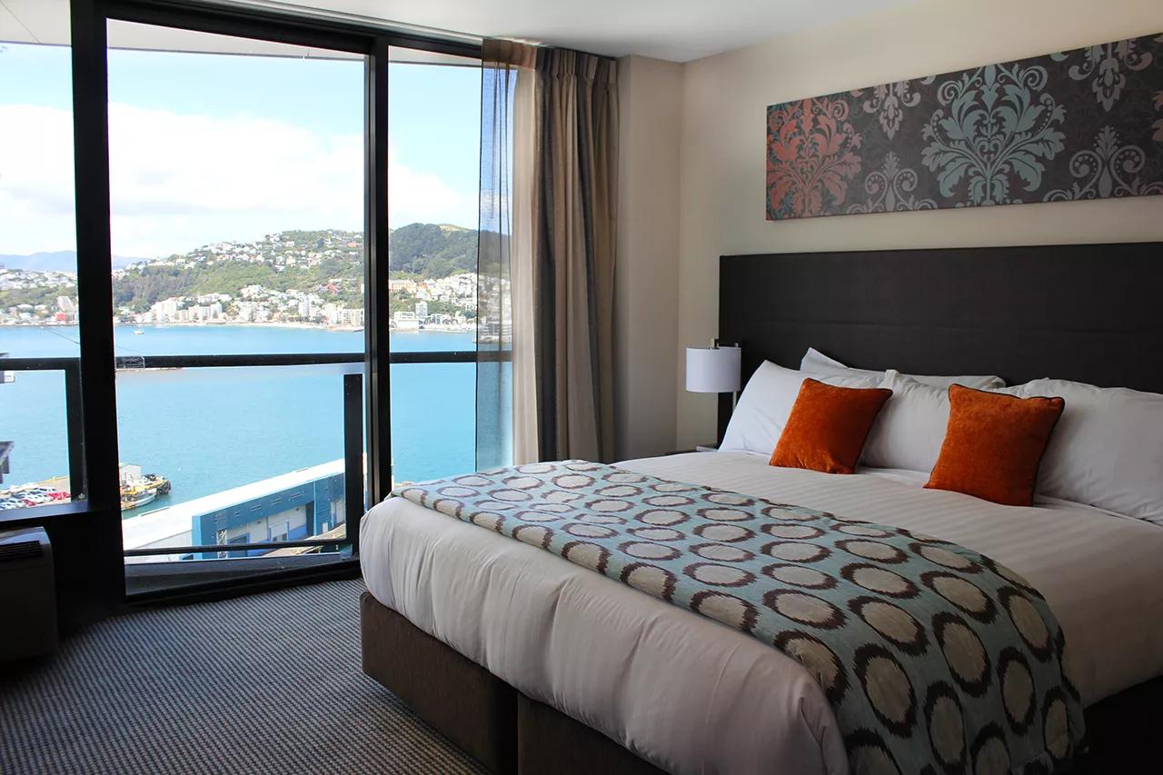 A room at the Rydges Hotel located at 75 Featherston Street, Pipitea, Wellington, with a king-sized bed, grey carpet, beige walls and a large window with a view of the harbour. 