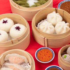 Close up of knitted yum cha items displayed in bamboo steamer baskets, on a red table.