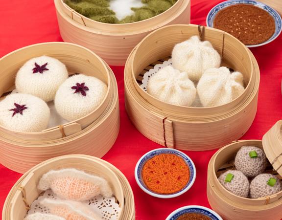 Close up of knitted yum cha items displayed in bamboo steamer baskets, on a red table.