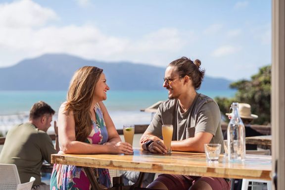 Two peple sitting outside at a bar enjoying drinks on a sunny day with Kapiti Island in the background.