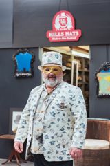 A person wearing a matching suit jacket and top hat that is off-white with a print of hats and a gold eye mask.